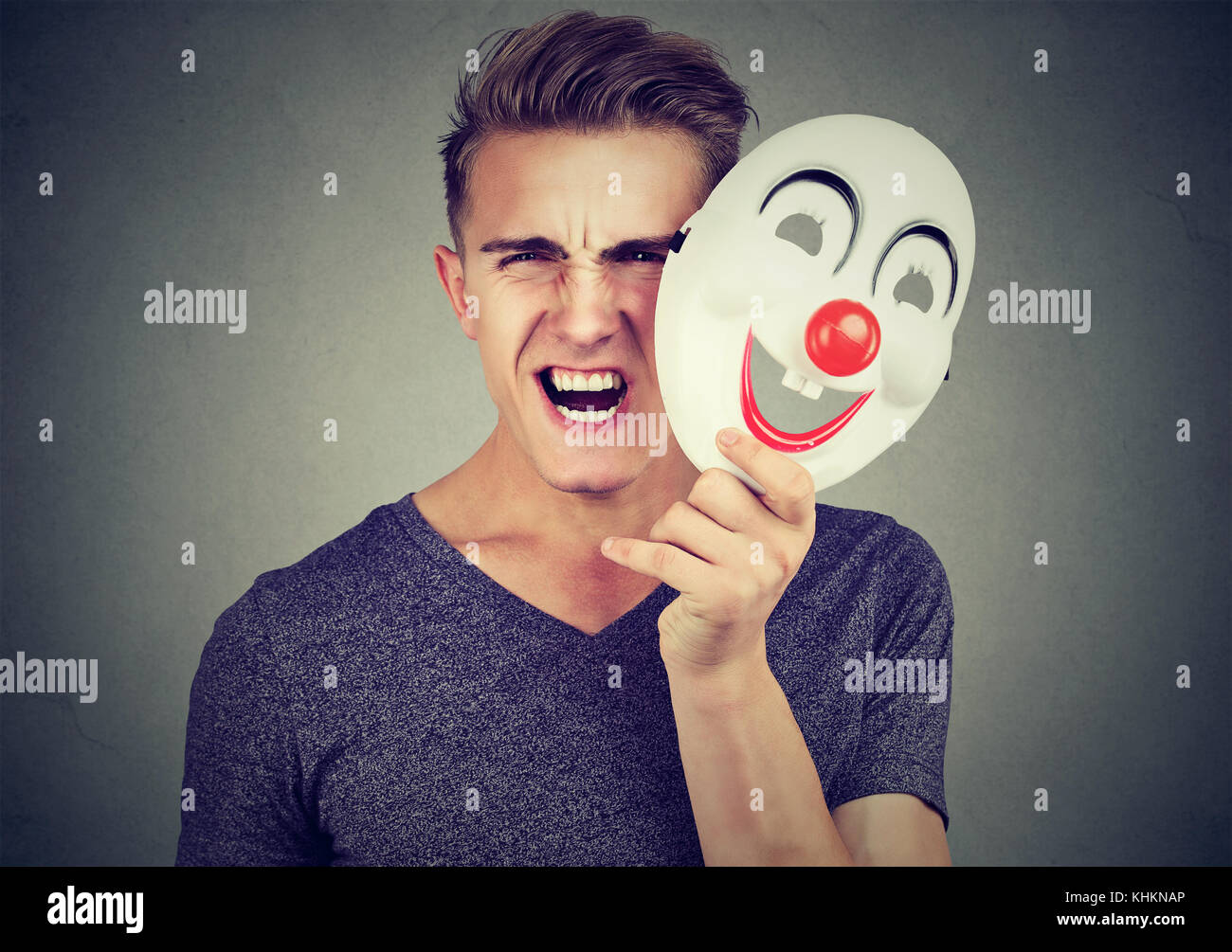 Ritratto arrabbiato urlando uomo togliersi la maschera di clown esprimendo felicità isolato su sfondo grigio. le emozioni umane sensazioni Foto Stock