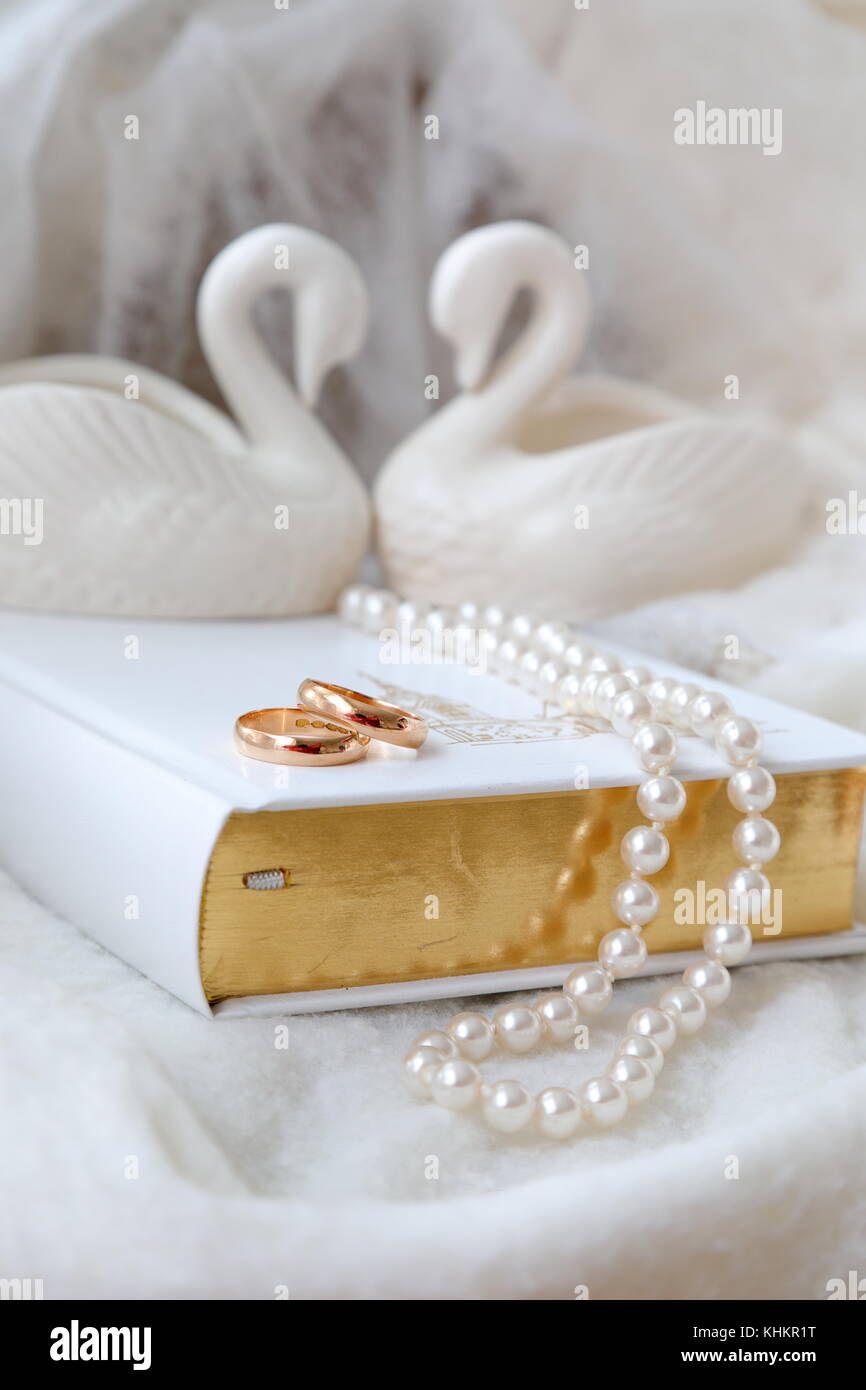 Due fedi nuziali d'oro sulla Bibbia bianca. Perle bianche e due cigni bianchi in porcellana. Sfondo bianco. Versione verticale della foto. Foto Stock