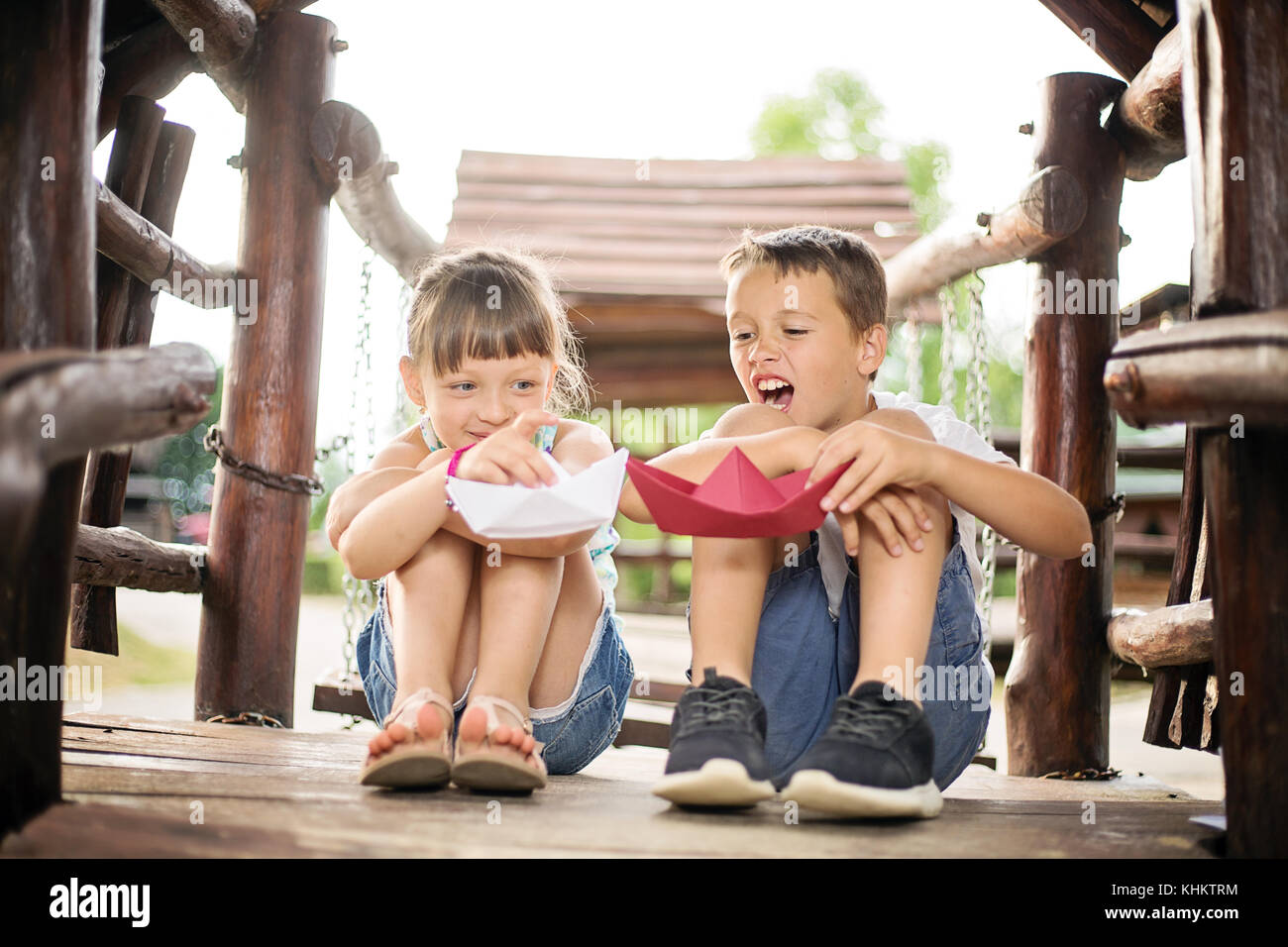 Due giovani bambini caucasici seduto accanto a ogni altro con le ginocchia piegate in una casa di legno, all'aperto o sulla soleggiata giornata estiva, giocando con barche di carta Foto Stock