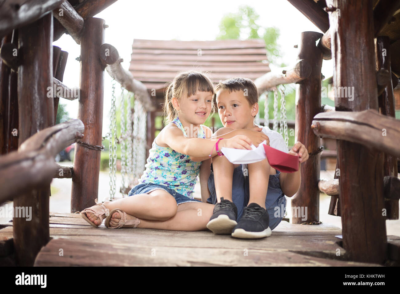 Due giovani bambini caucasici seduto accanto a ogni altro con le ginocchia piegate in una casa di legno, all'aperto o sulla soleggiata giornata estiva, giocando con barche di carta Foto Stock
