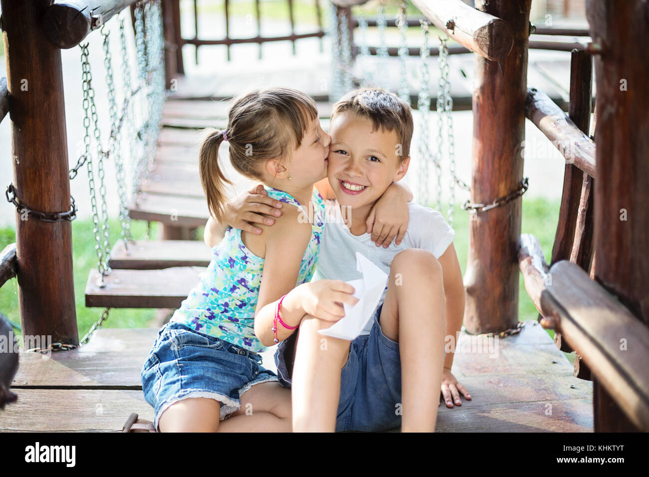 Due giovani caucasici abbracciando bambini e una ragazza sta baciando un ragazzo su una guancia. Essi sono seduti in una casa di legno in un parco giochi per bambini al giorno d'estate e di sole. Foto Stock
