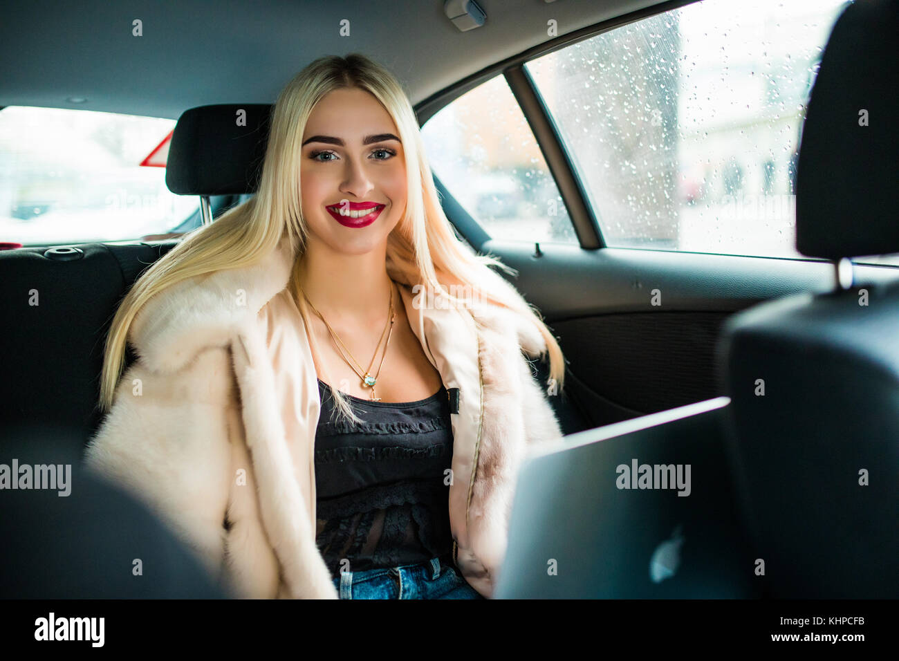 Bella donna di occhiali è utilizzando un computer portatile e sorridenti mentre è seduto sul sedile posteriore in auto Foto Stock