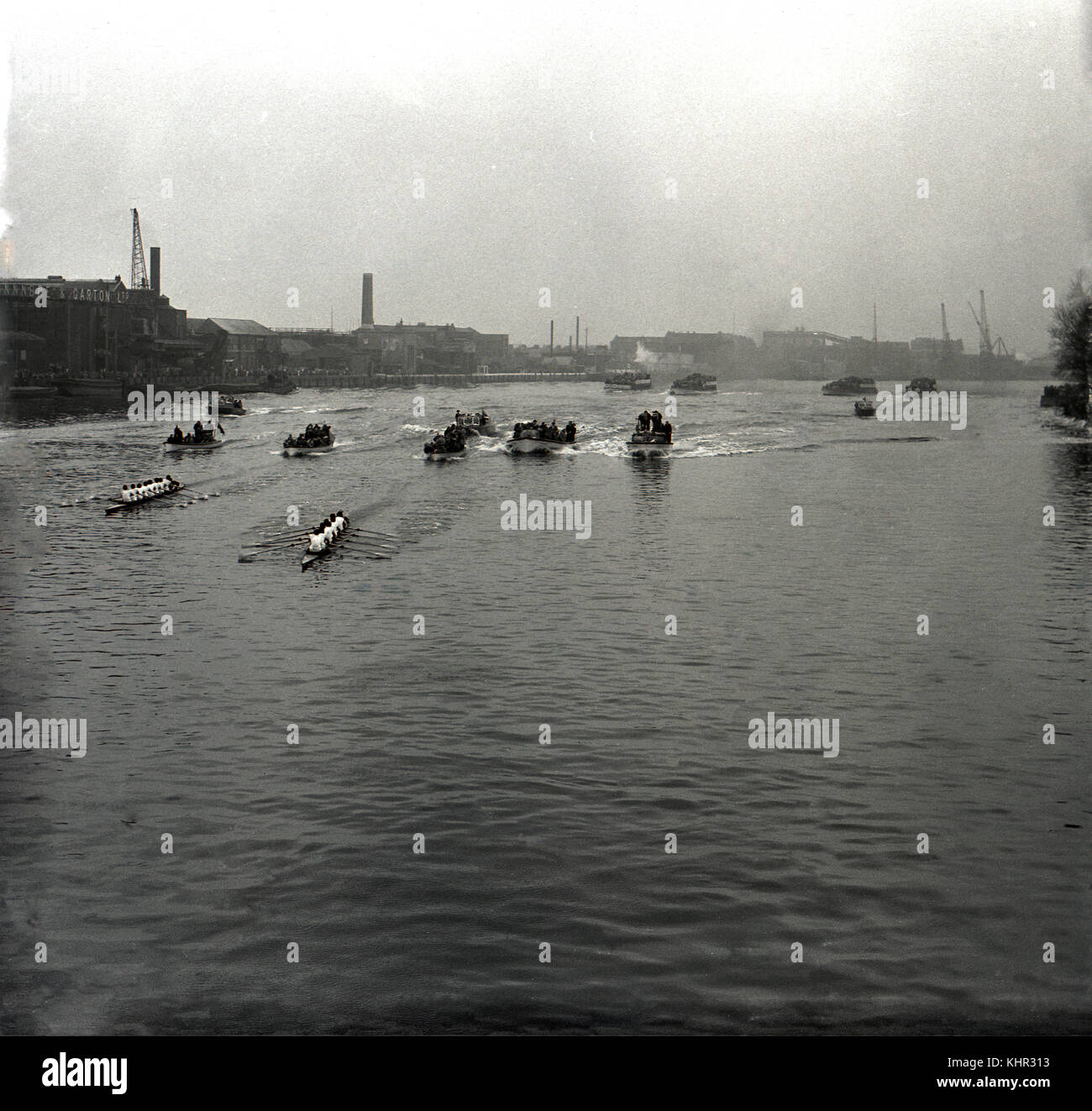 Degli anni Cinquanta, historial immagine che mostra le barche e la flottiglia della famosa università gara di canottaggio, Oxford e Cambridge Boat Race scendendo lungo il corso del fiume Tamigi passato il terrapieno di Hammersmith, Londra, Inghilterra. Sulla sinistra, i numerosi pontili industriali di questo tempo, compreso il Manbre & Garton magazzino zucchero. Foto Stock