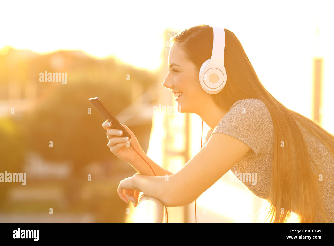 Vista laterale verticale di una ragazza felice ascolto di musica con cuffie e smart phone in una casa balcone al tramonto con una luce arancione Foto Stock