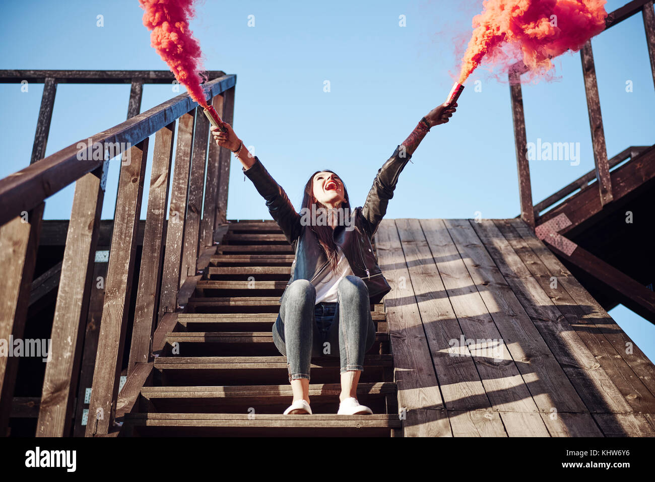 Giovane donna seduta sui gradini in legno, tenendo Razzi manuali, rosso fumo versando da flares, basso angolo di visione Foto Stock
