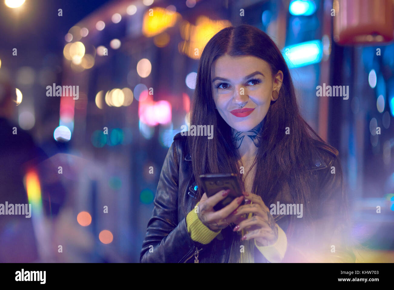 Giovane donna all'aperto durante la notte, tenendo lo smartphone, tatuaggi a portata di mano e collo Foto Stock