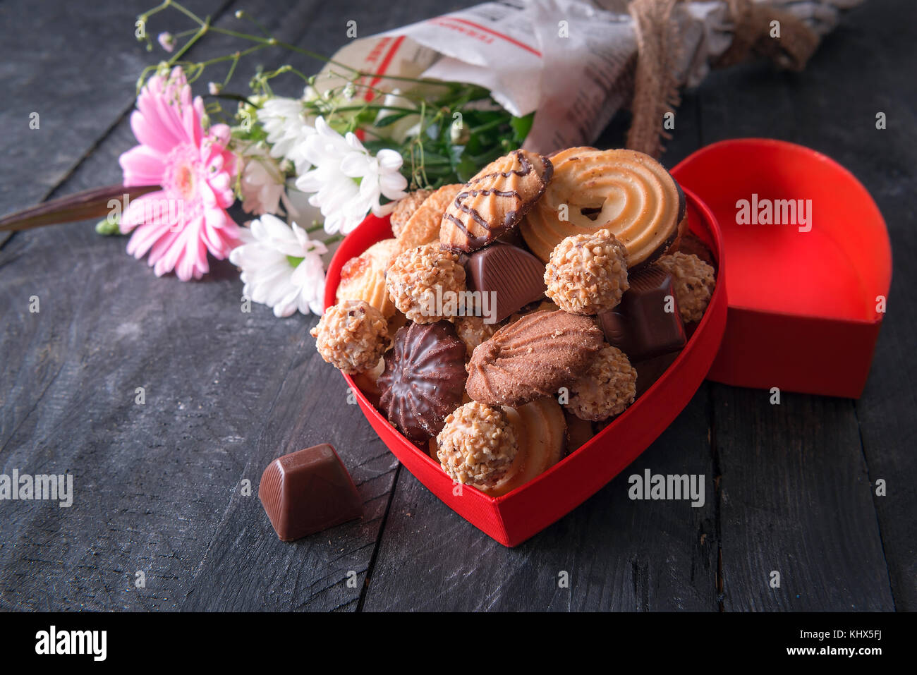 Biglietto di auguri idea con una a forma di cuore scatola piena di gustosi biscotti, cioccolatini e un mazzo di fiori avvolti in un giornale in background. Foto Stock