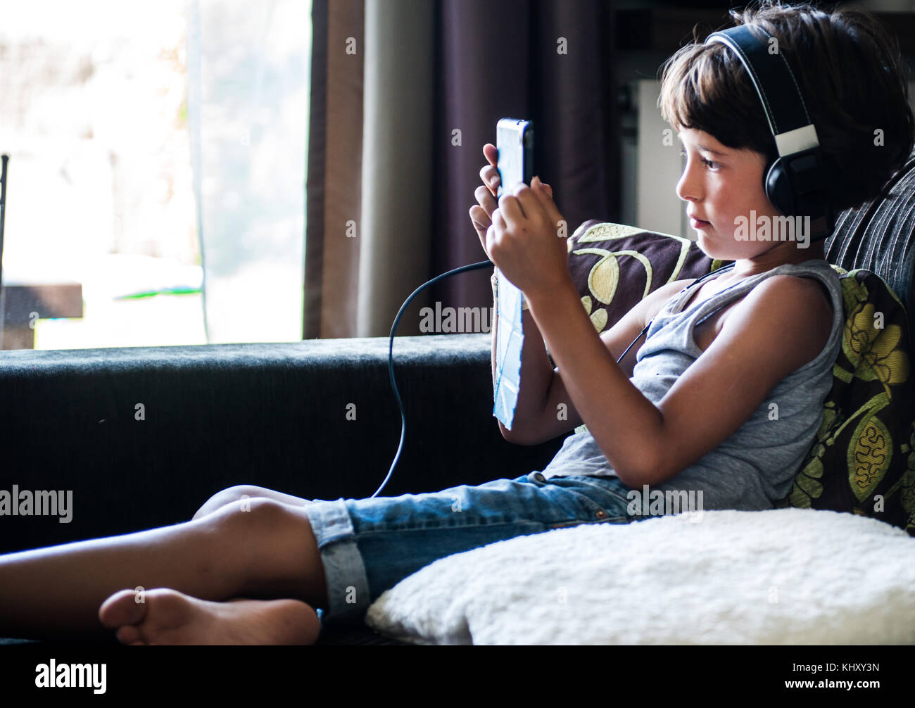 Ragazzo seduto sul divano ascoltando le cuffie e fissando a tavoletta digitale Foto Stock