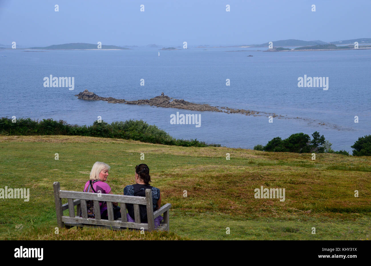 Due donne sulla panca in legno,parlando e ammirando la vista vicino halangy village & sepoltura camera sull'isola di St Marys nelle isole Scilly, UK. Foto Stock
