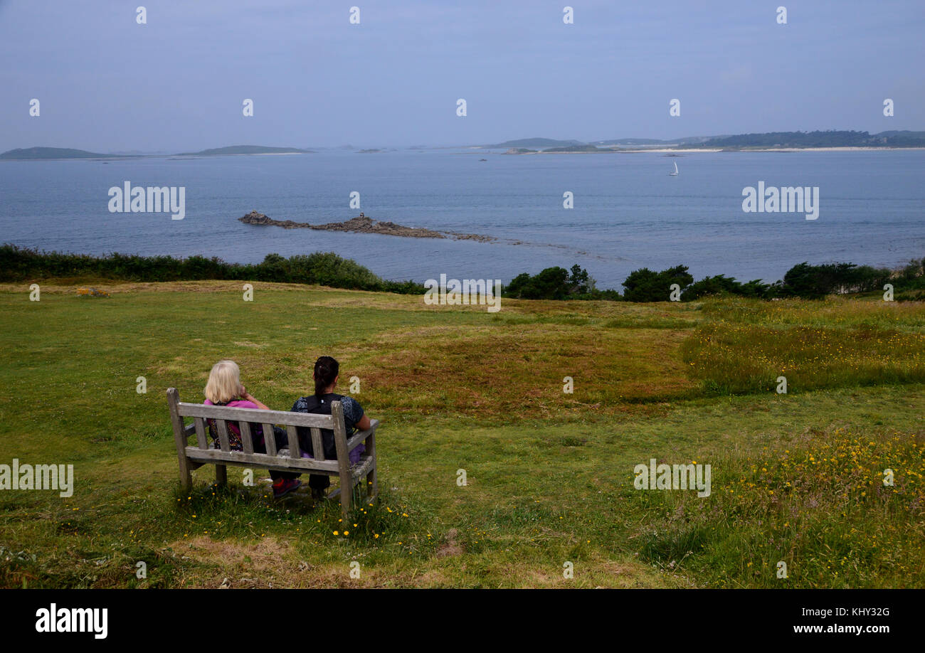 Due donne sulla panca in legno,parlando e ammirando la vista vicino halangy village & sepoltura camera sull'isola di St Marys nelle isole Scilly, UK. Foto Stock