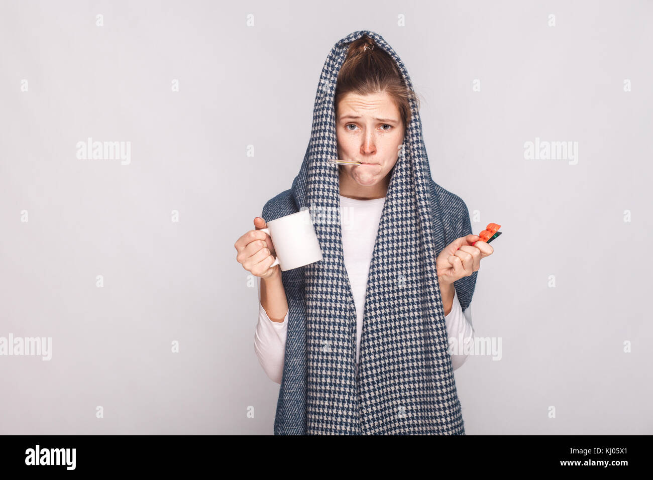 Giovane donna con il grigio sciarpa, coppa di ritegno, termometro e pillole. interna shot. Foto Stock