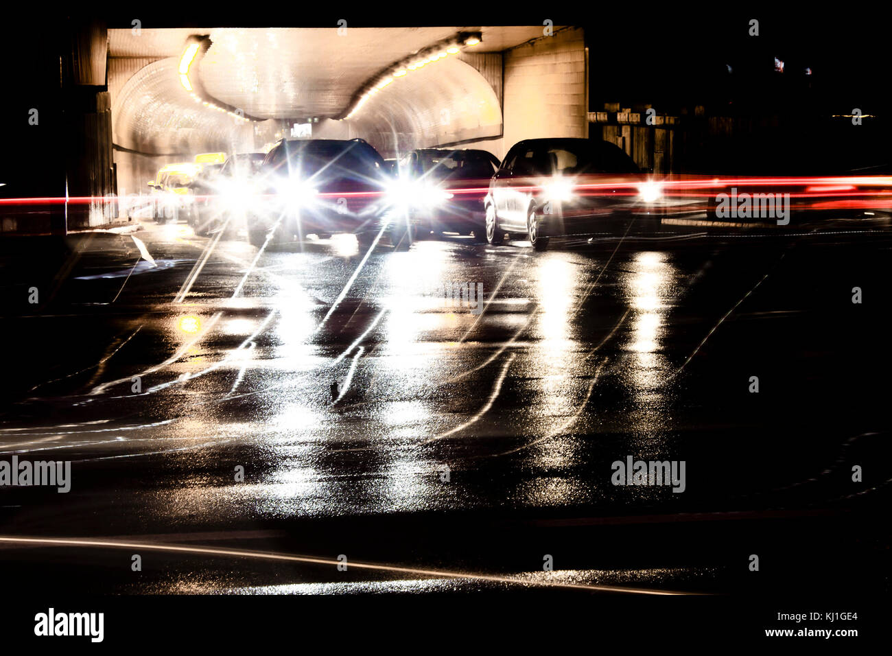 Sfocato il traffico notturno di pioggia per le strade delle città. vetture accodate all'uscita del tunnel in attesa presso l'intersezione durante la guida di veicoli che si spostano oltre in motion blur. Foto Stock