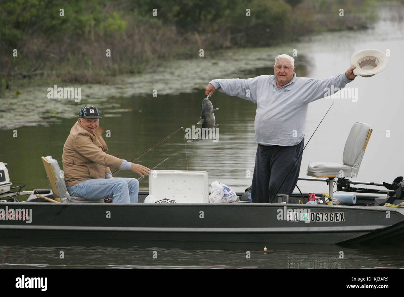 Due uomini anziani godendo la pesca dalla barca per un uomo è in piedi e sollevando hat mentre mostra il pesce appena pescato Foto Stock