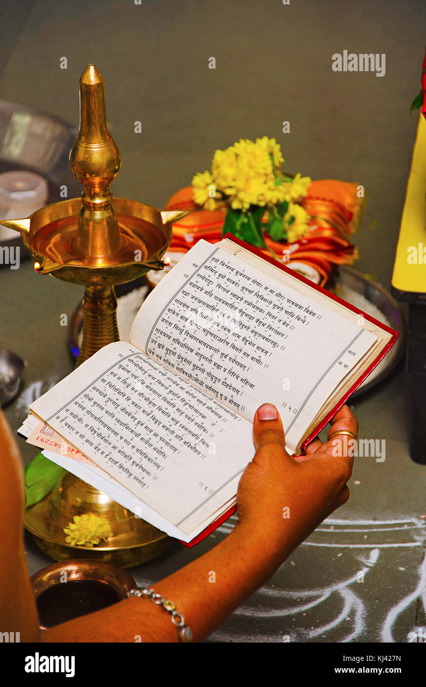 Sacerdote tenendo un libro contenente i rituali nella sua mano destra con una tradizionale lampada, fiori e rangoli in background, Belgavi, Karnataka, India Foto Stock