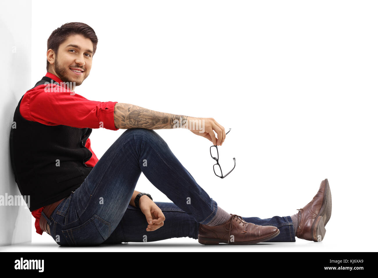 Tatuato ragazzo seduto sul pavimento appoggiato contro una parete e guardando la telecamera isolata su sfondo bianco Foto Stock