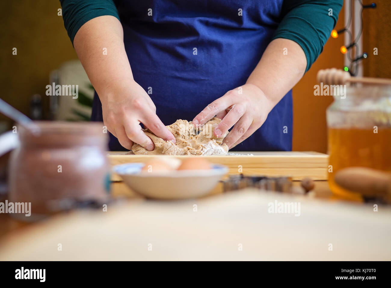 Close up della giovane donna mani impastare la pasta su una tavola di legno. ella è in piedi in cucina, dietro un tavolo accanto a una finestra decorata con chri Foto Stock