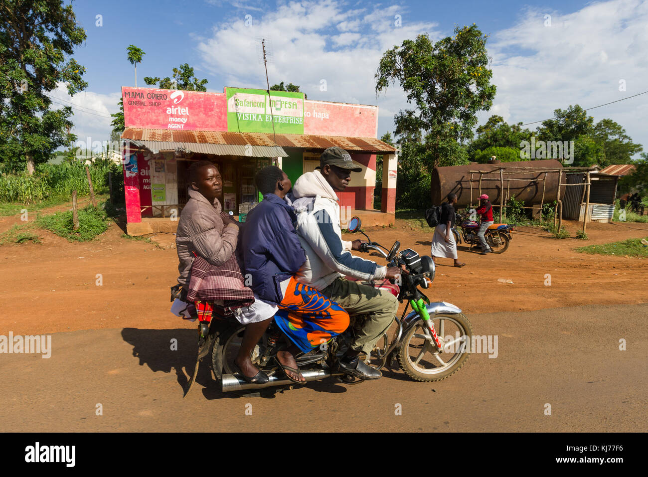 Un boda boda moto taxi driver con due unità di passeggeri su strada in passato una piccola fila di negozi, Kenya, Africa orientale Foto Stock