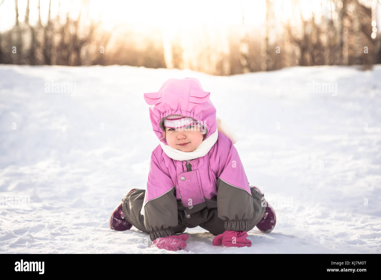 Allegro baby a carponi giocando sulla neve nel freddo inverno pieno di sole giorno in vestiti caldi con la luce del sole attraverso gli alberi Foto Stock