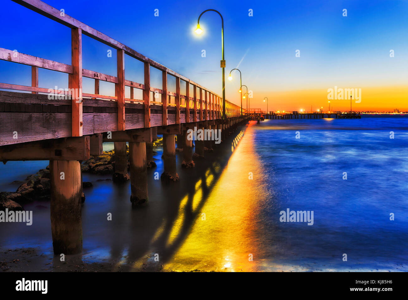 Dark tramonto sul centro storico pontile in legno a Port Melbourne sulla spiaggia della Baia di Port Phillip. Le luci di strada e il sole di setting riflettono in sfocata acque blu. Foto Stock