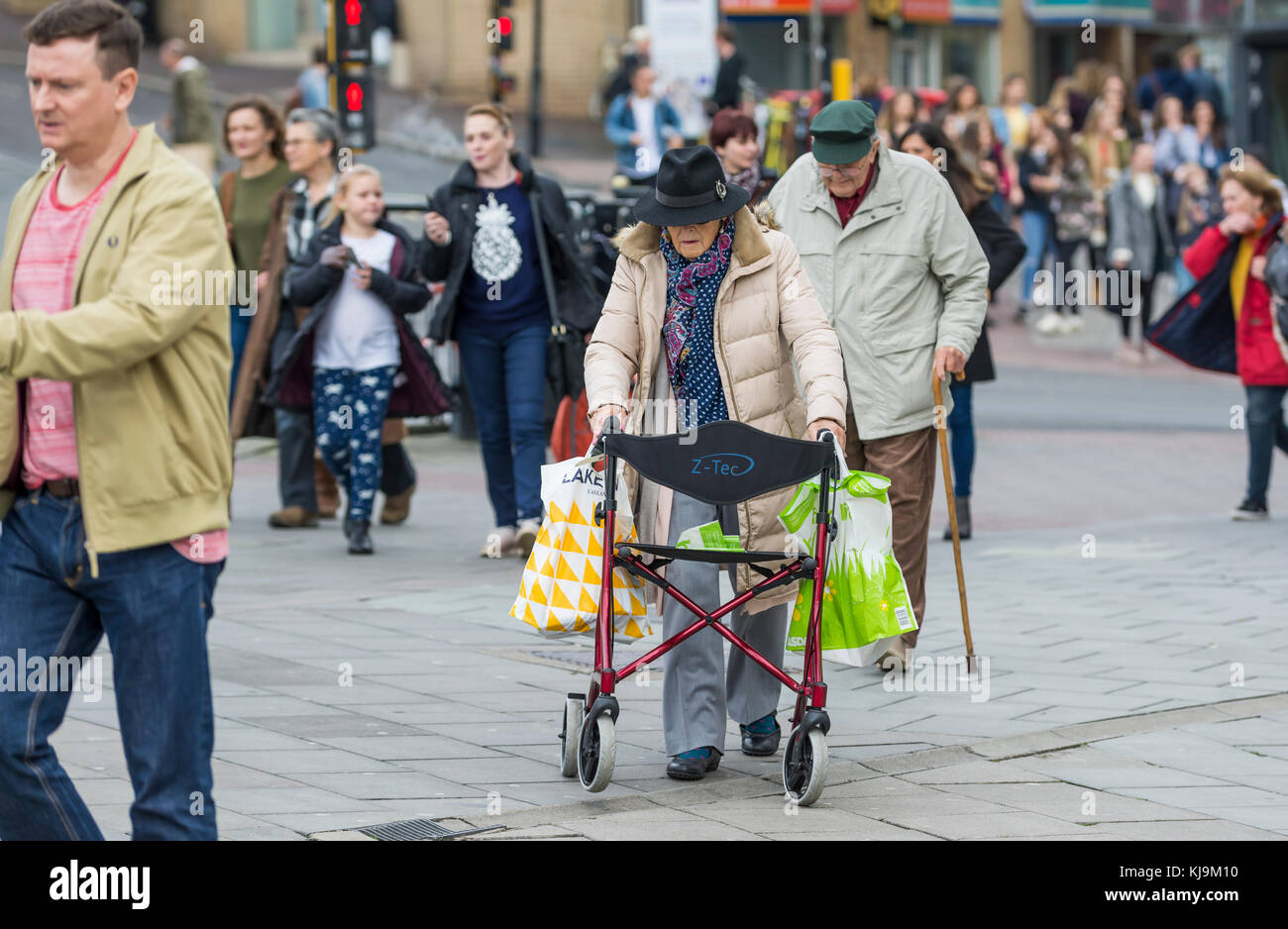 Signora anziana a piedi attraverso una trafficata città popolata con l aiuto di un gommato Zimmer frame in Inghilterra, Regno Unito. Donna anziana con un Rollator aiuti a piedi. Foto Stock