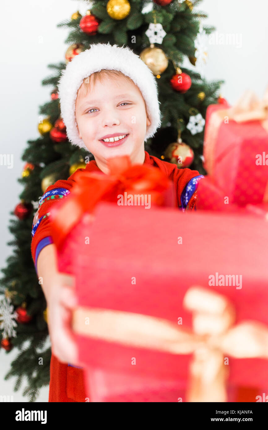 Closeup ritratto di sorridenti bambino holding pila di scatole presenti nelle mani di. ragazzo indossa santa hat e pigiami rosso. vacanze albero verde in backgroun Foto Stock