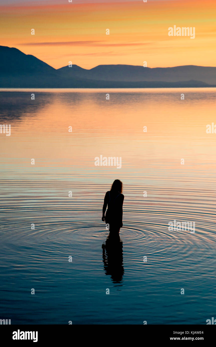 Una donna sola bagnante viene riflessa come Ella entra nella calme acque del mare Egeo. Ella si stagliano i colori fiammeggianti di un mattino cielo. Foto Stock