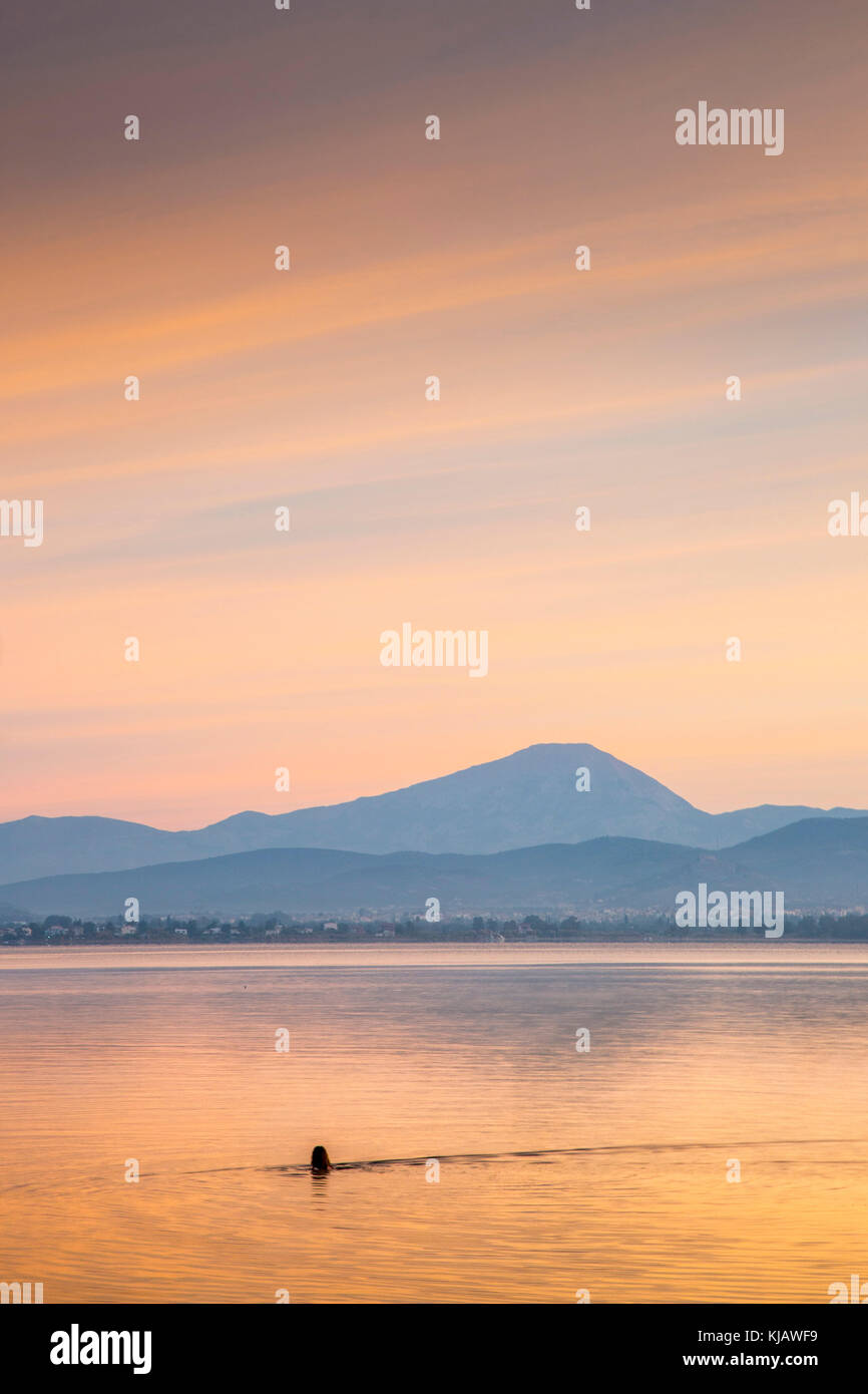 Il colore arancio alba sky è riflessa nel mare calmo come un bagnante solitario nuota nella parte anteriore del monte Olimpo in Grecia. Foto Stock