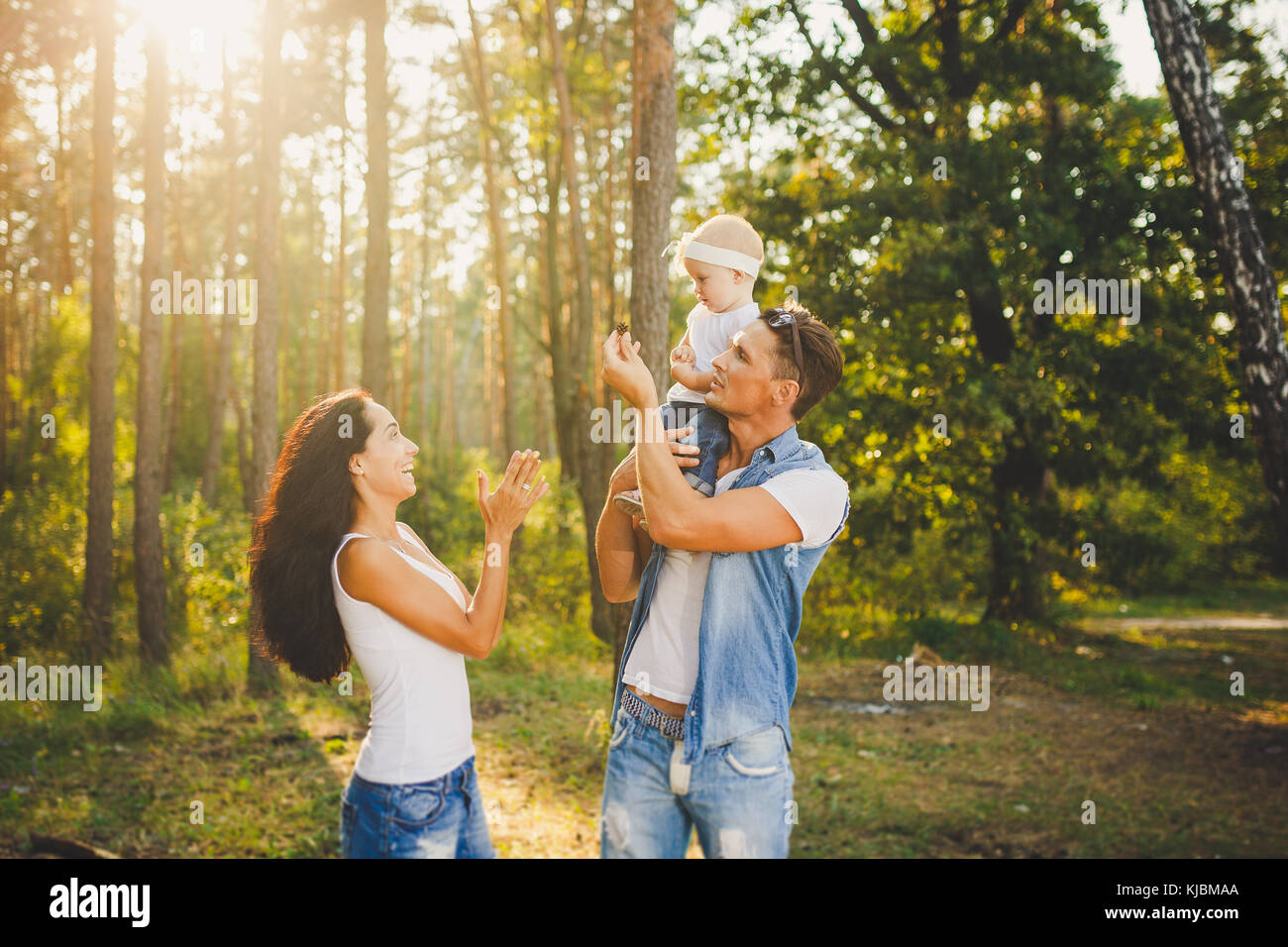 Elegante giovane famiglia mamma, papà e la figlia di un anno di età bionda seduta con il padre sulle spalle giocando felice e sorridente, all'esterno al di fuori della città nei boschi in estate al tramonto. Foto Stock