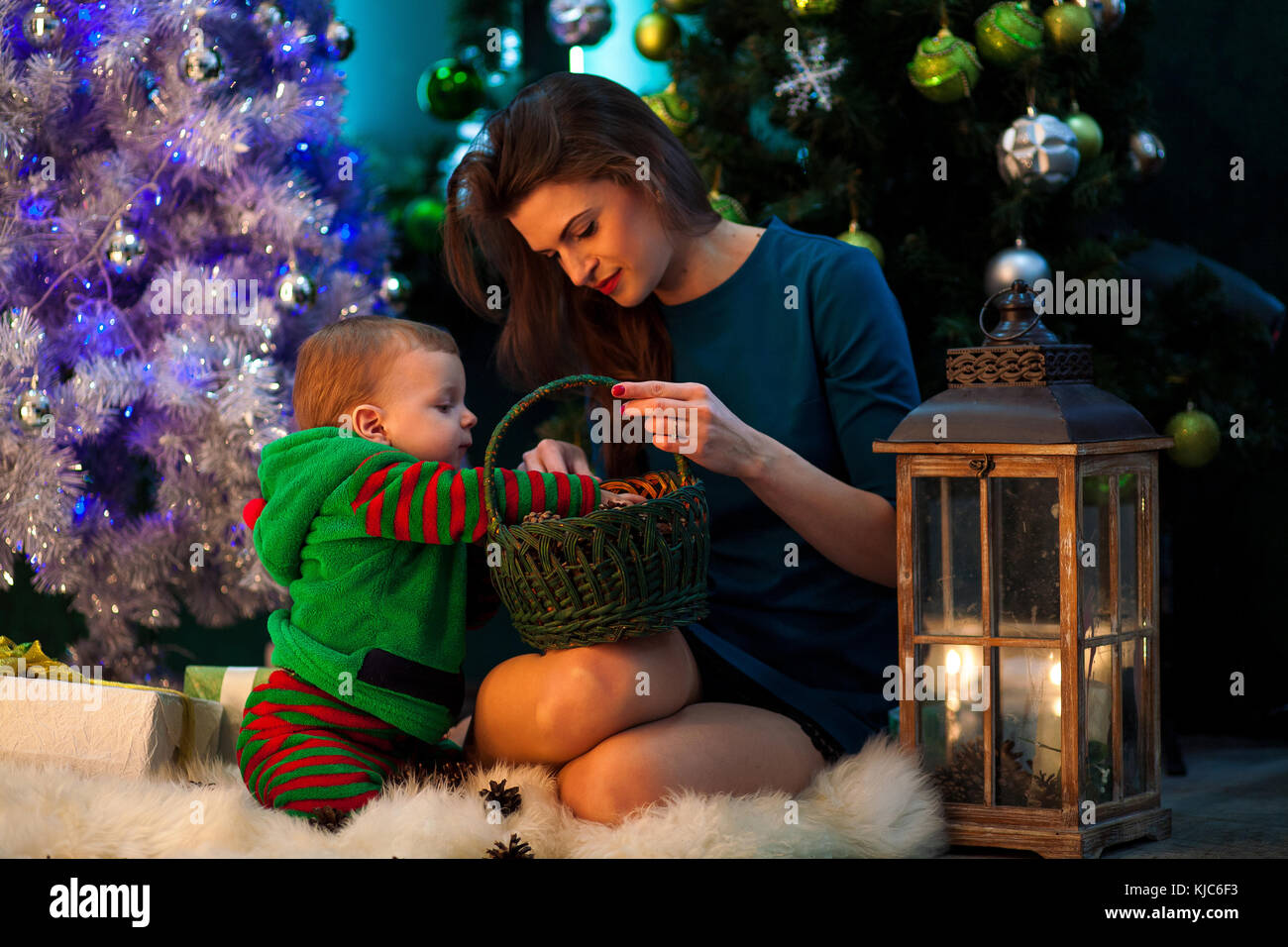 Felice madre con il suo piccolo figlio di sedersi sul bianco tappeto di pelliccia e guardare i regali di Natale nel cesto in vimini su sfondo dell albero di natale, decorazioni e li Foto Stock