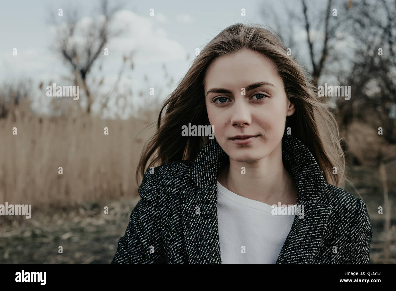 Bella e giovane donna con capelli lunghi biondi, ampia sopracciglia, tutte le labbra, occhi blu vestiti di lana cappotto. Ragazza guardando la fotocamera. Giunco di palude di sfondo. Foto Stock