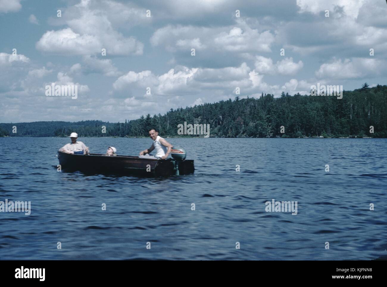 Una fotografia di tre uomini in una barca sul lago, un uomo si siede nella parte posteriore della barca per utilizzare il motore fuoribordo mentre un altro uomo si siede frontalmente il terzo uomo è seduto sul pavimento della barca in modo che solo la sua testa è visibile, il lago può essere visto in allungamento della distanza, è circondato da alberi di alto fusto e le colline ondulate, 1982. Foto Stock