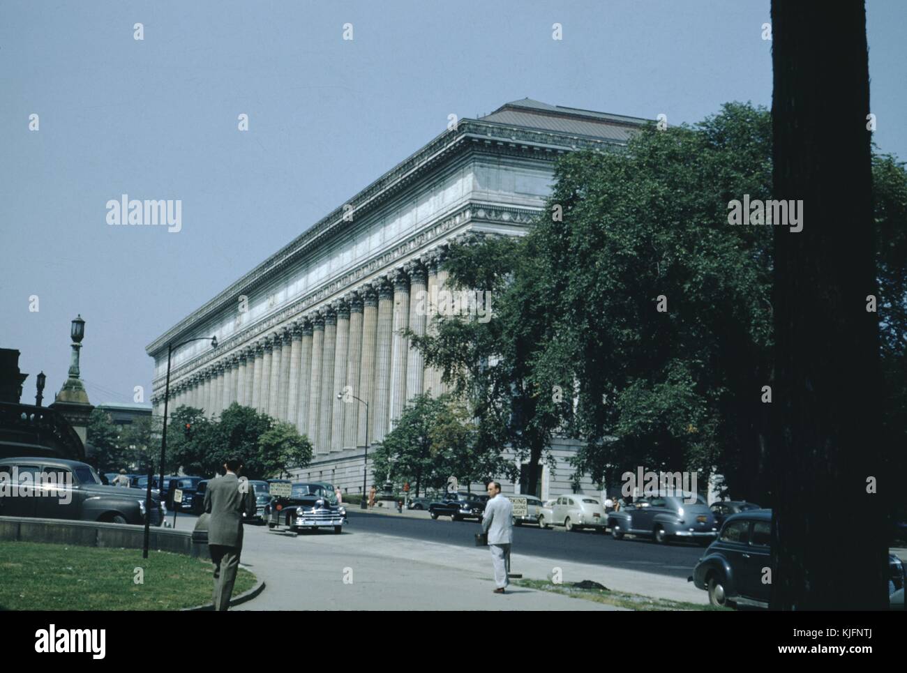 Una fotografia contenente una vista di tre quarti di un edificio con molte colonne corinzie, due uomini in tuta camminano verso l'edificio, più di una dozzina di auto d'epoca possono essere viste parcheggiate lungo la strada, gli alberi possono essere visti crescere dai marciapiedi nell'area intorno all'edificio, 1952. Foto Stock