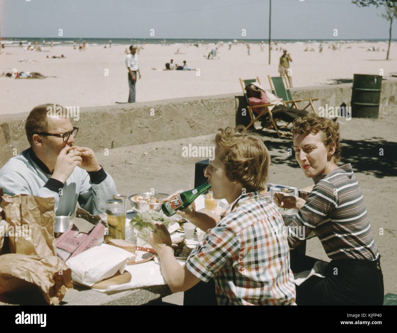 Tre persone, un uomo e due donne, sedute a un tavolo, su un sentiero di cemento vicino alla spiaggia, mangiando una grande quantità di cibo e bevendo una bibita, sullo sfondo, persone sdraiate sulla sabbia in spiaggia, 1952. Foto Stock