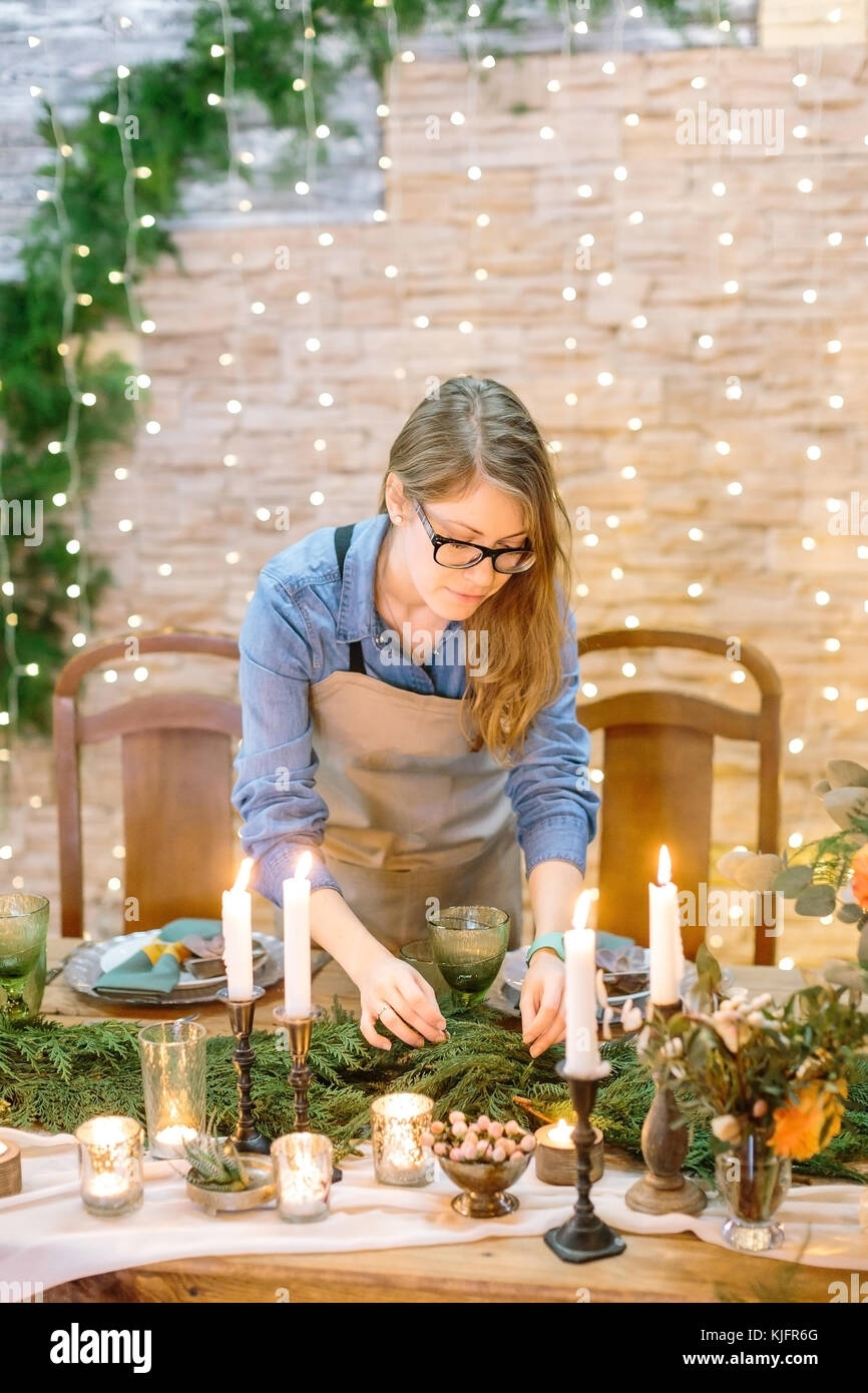 Piante, design, parte il concetto di spazio. ragazza caucasica ondulata con capelli biondi rendendo il design floreale della tabella, decorare con rami di conifere, indossa gli occhiali e semplici ma adorabile maglietta denim Foto Stock