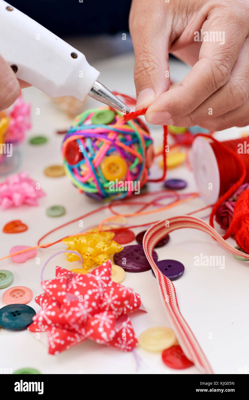 Primo piano di un giovane uomo che fa un Natale fatti a mano palla usando una pistola per colla a caldo con le stringhe e i pulsanti di colori diversi e una pila di differenti hab Foto Stock