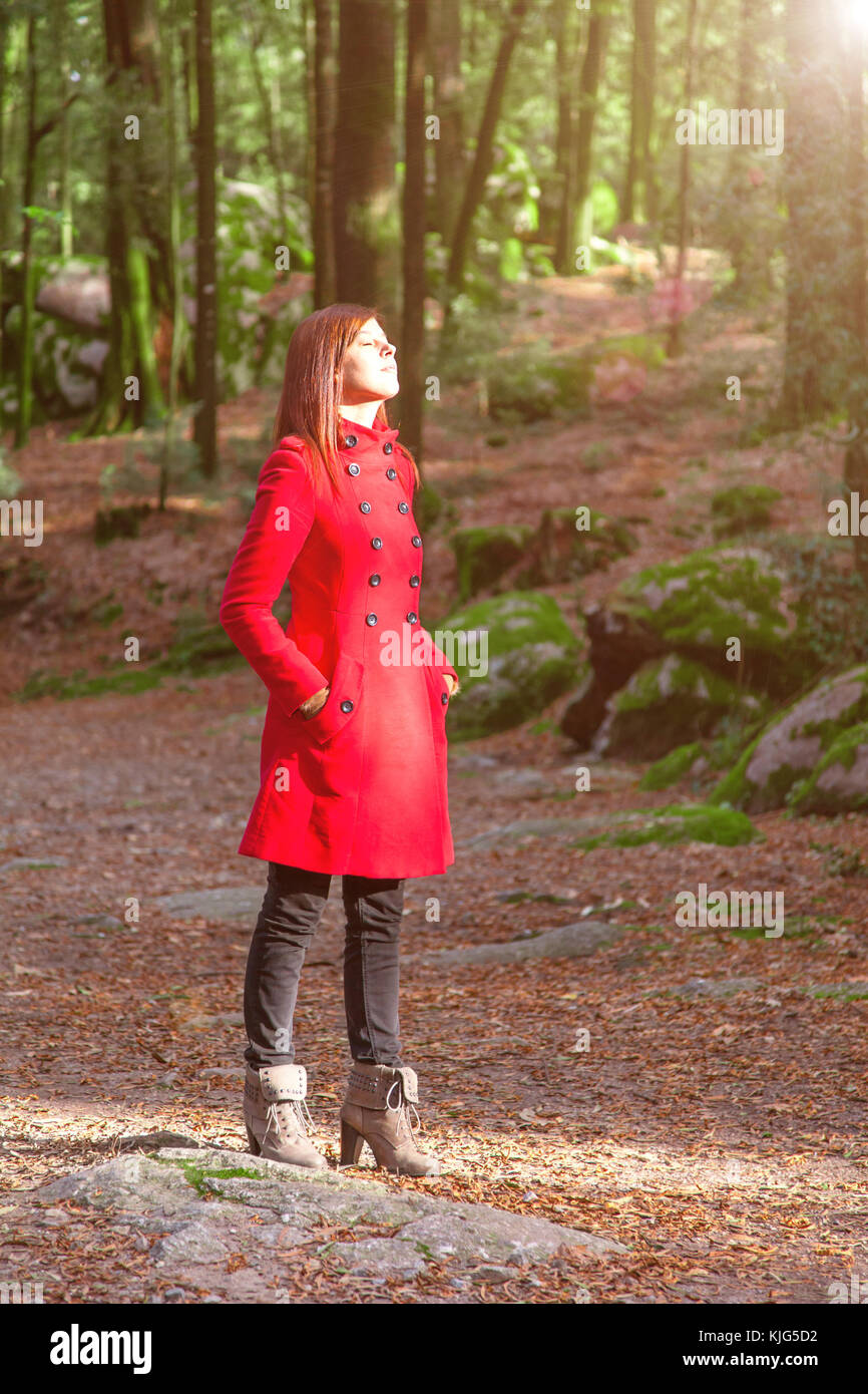 Donna godendo il calore del sole d'inverno solo su di un parco forestale percorso indossando un soprabito rosso Foto Stock