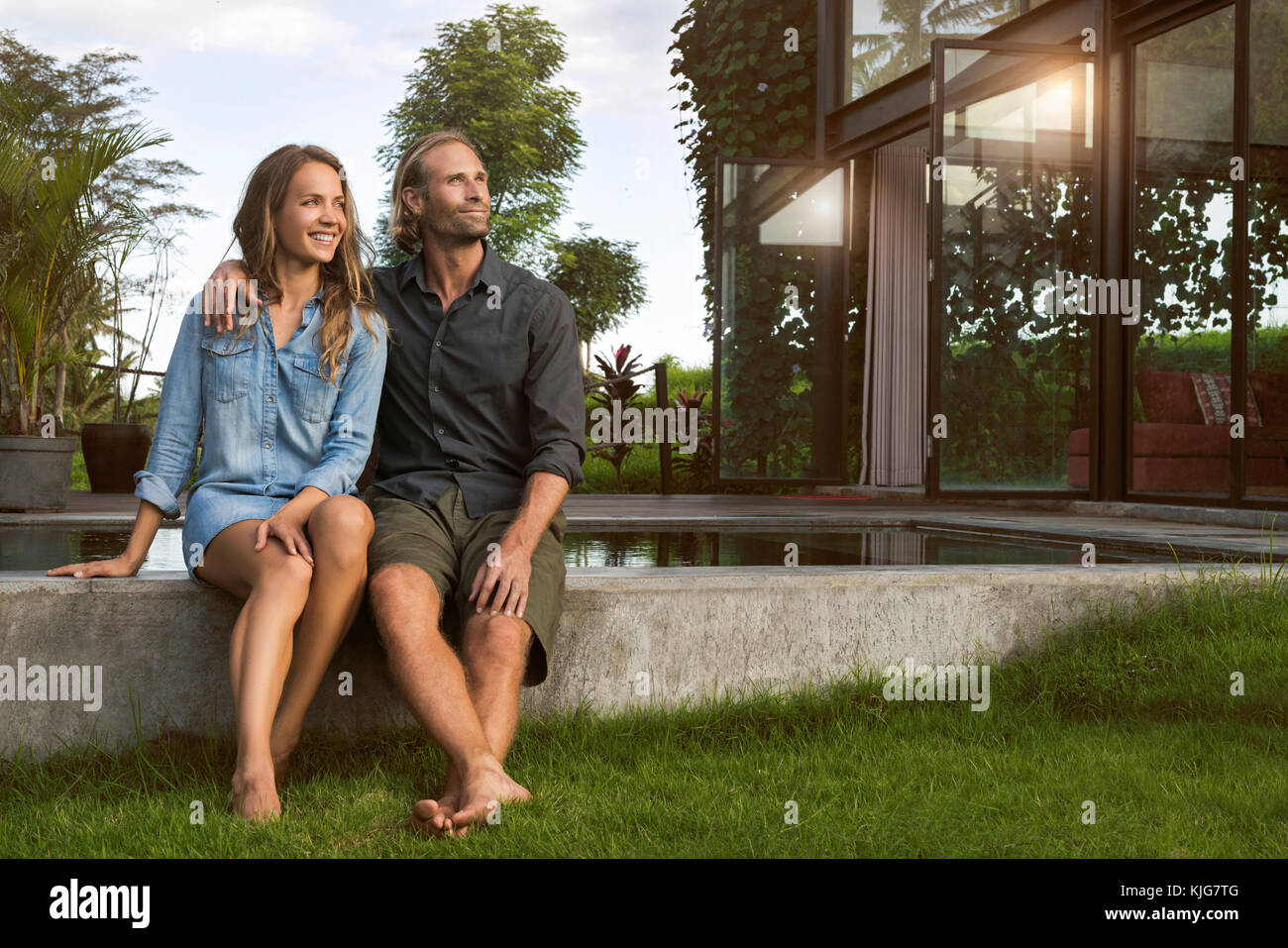 Sorridente abbracciando giovane seduto sul bordo di una piscina di fronte alla loro casa di design Foto Stock