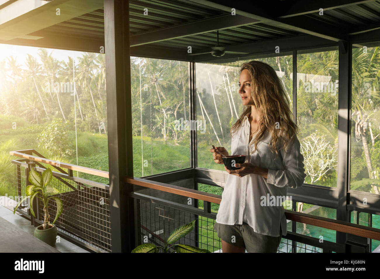 Donna sorridente mangiare cereali in design contemporaneo casa con piscina e la facciata di vetro circondato da un lussureggiante giardino tropicale Foto Stock