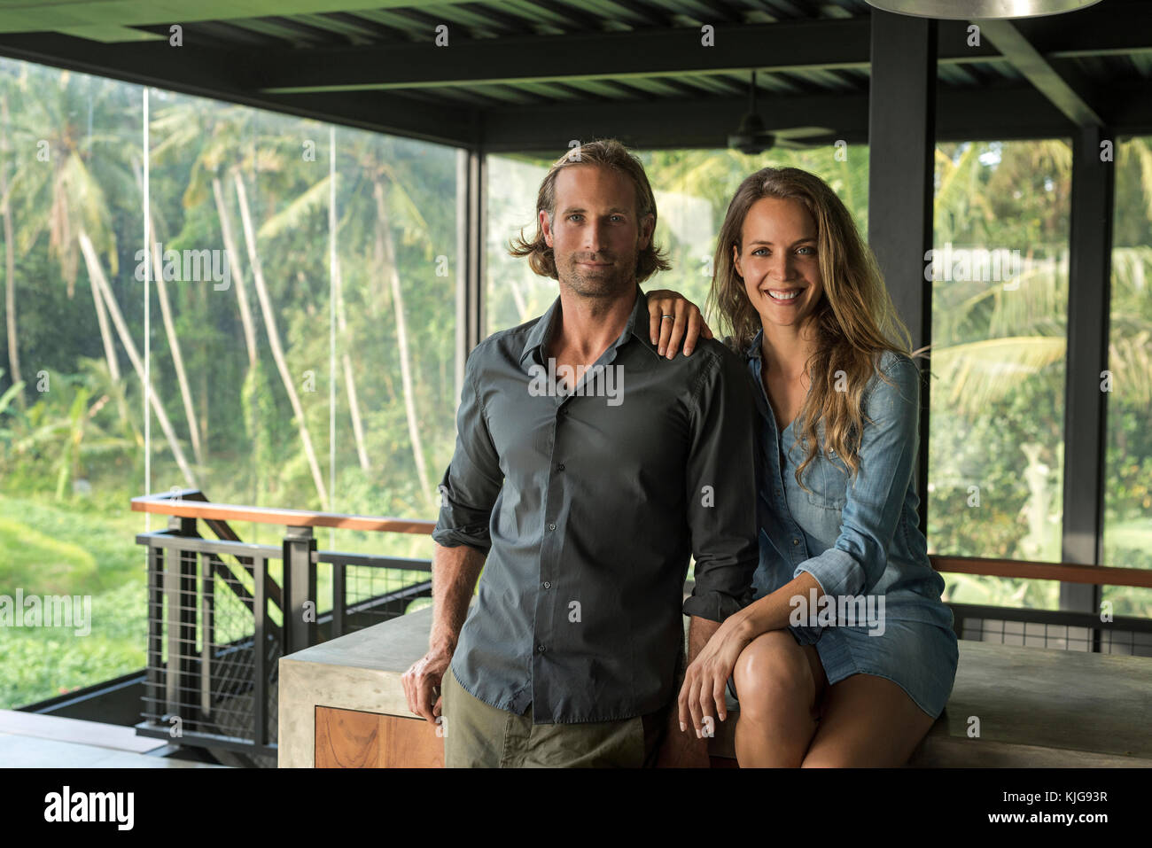 Coppia sorridente in telecamera in design contemporaneo casa con facciata di vetro circondato da un lussureggiante giardino tropicale Foto Stock
