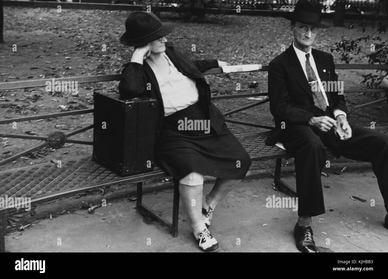 Una fotografia di un uomo e di una donna seduti su una panchina di metallo, la donna indossa una giacca, una gonna, e cappello, lei è appoggiata su una grande cassa nera, l'uomo indossa un vestito e una cravatta insieme a un cappello, le foglie possono essere viste coprire il terreno a Jackson Square dietro di loro, New Orleans, Louisiana, 1935. Dalla New York Public Library. Foto Stock