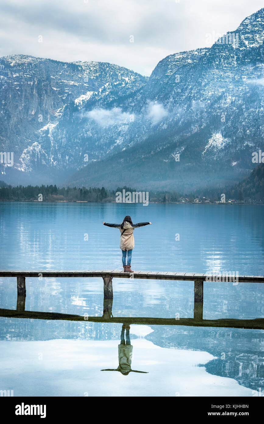 La ragazza con le braccia aperte, in piedi su uno stretto ponte di legno, godendo la vista delle Alpi austriache montagne riflesse nel lago hallstatter, Austria Foto Stock