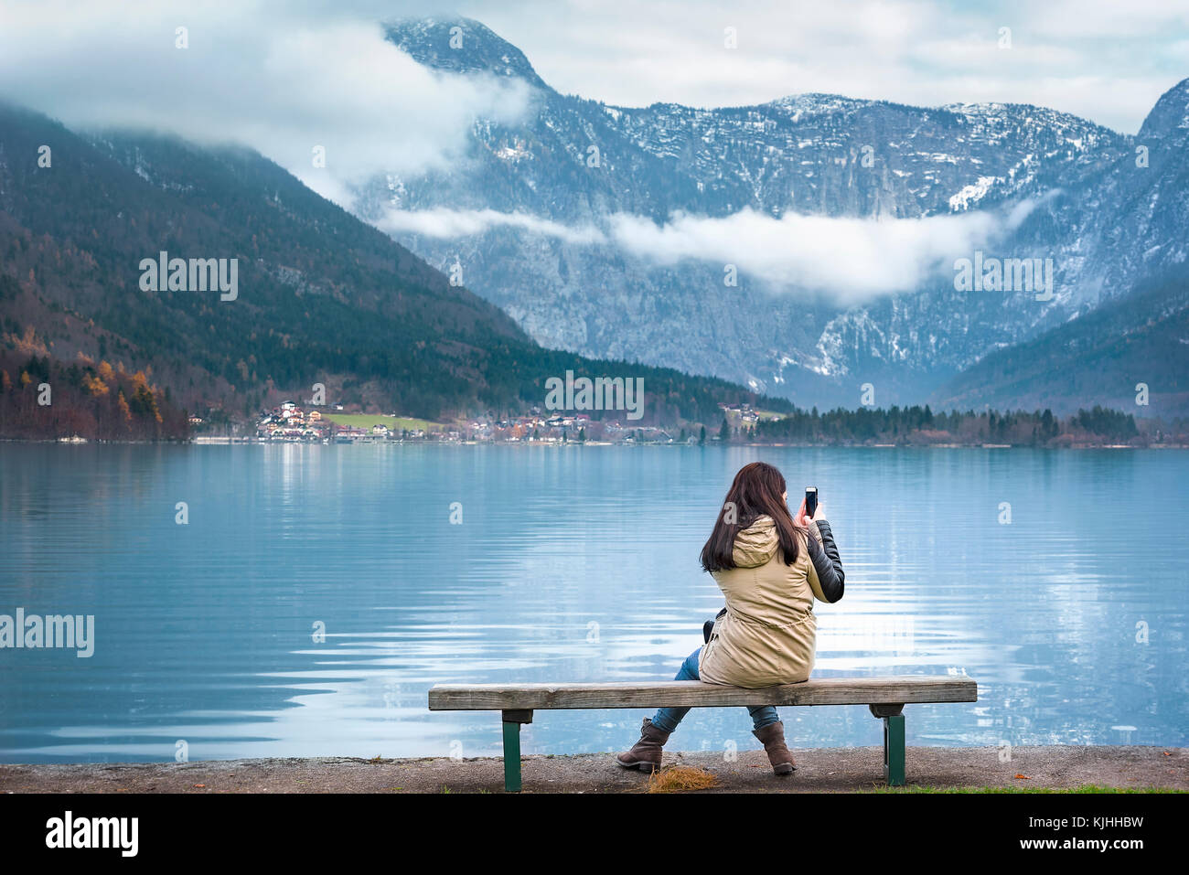 Donna seduta su una panca in legno, sulle rive del lago hallstatter, utilizza lo smartphone per fotografare le montagne austriache, di Hallstatt, Austria Foto Stock