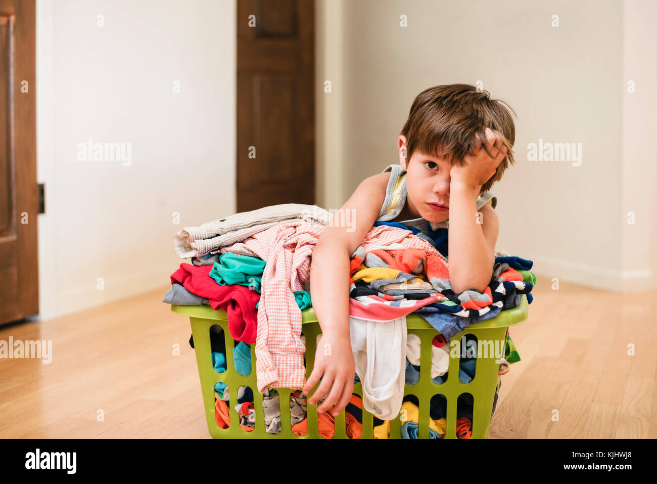 Ragazzo seduto sul pavimento appoggiandosi a un servizio lavanderia basked riempito con vestiti Foto Stock