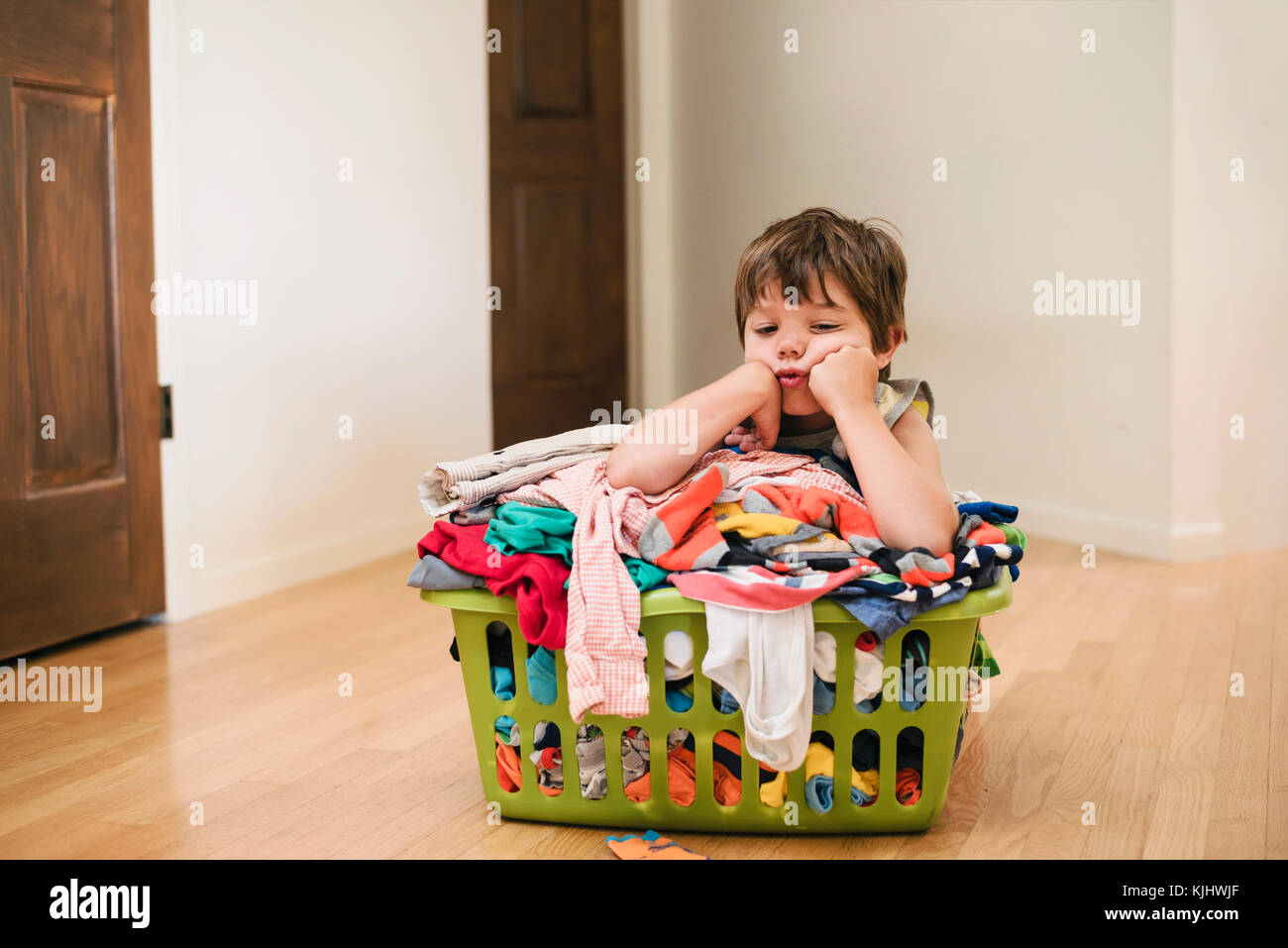 Stanco ragazzo seduto sul pavimento appoggiandosi a un servizio lavanderia basked riempito con vestiti Foto Stock