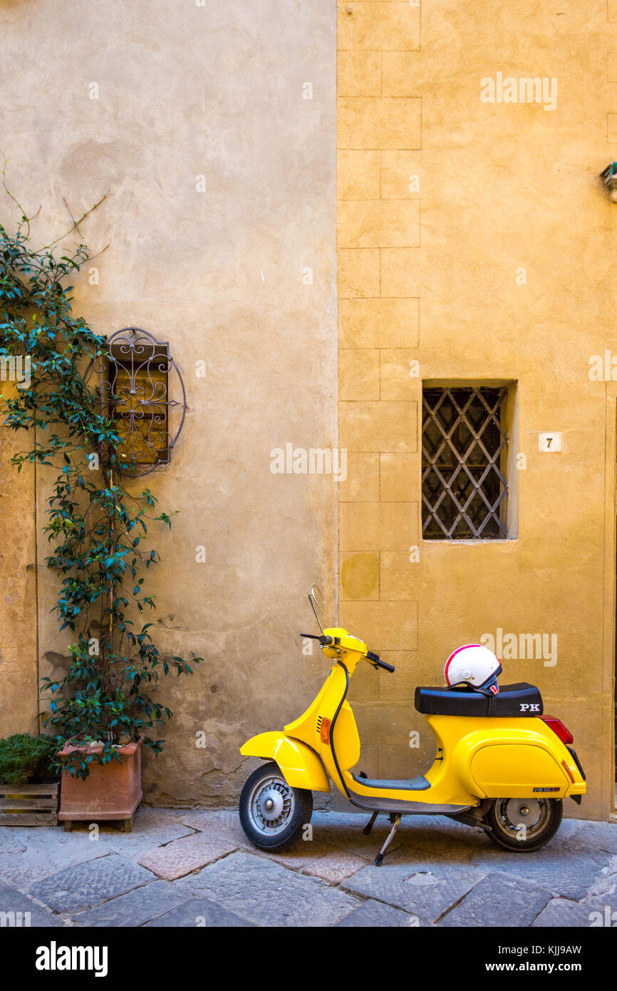 Vespa gialla in stile vintage con casco sul sedile, parcheggiata di fronte a una parete in pietra gialla in due tonalità presa nel villaggio di Pienza in Toscana. Nessuna gente Foto Stock