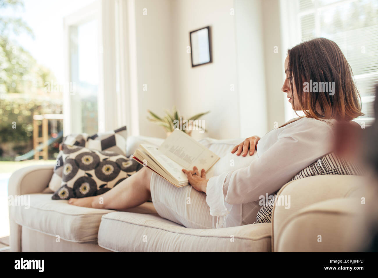 donna incinta seduta sul divano toccando la pancia e leggendo un libro. Aspettarsi che la madre si rilassasse sul divano preparasse un libro. Foto Stock