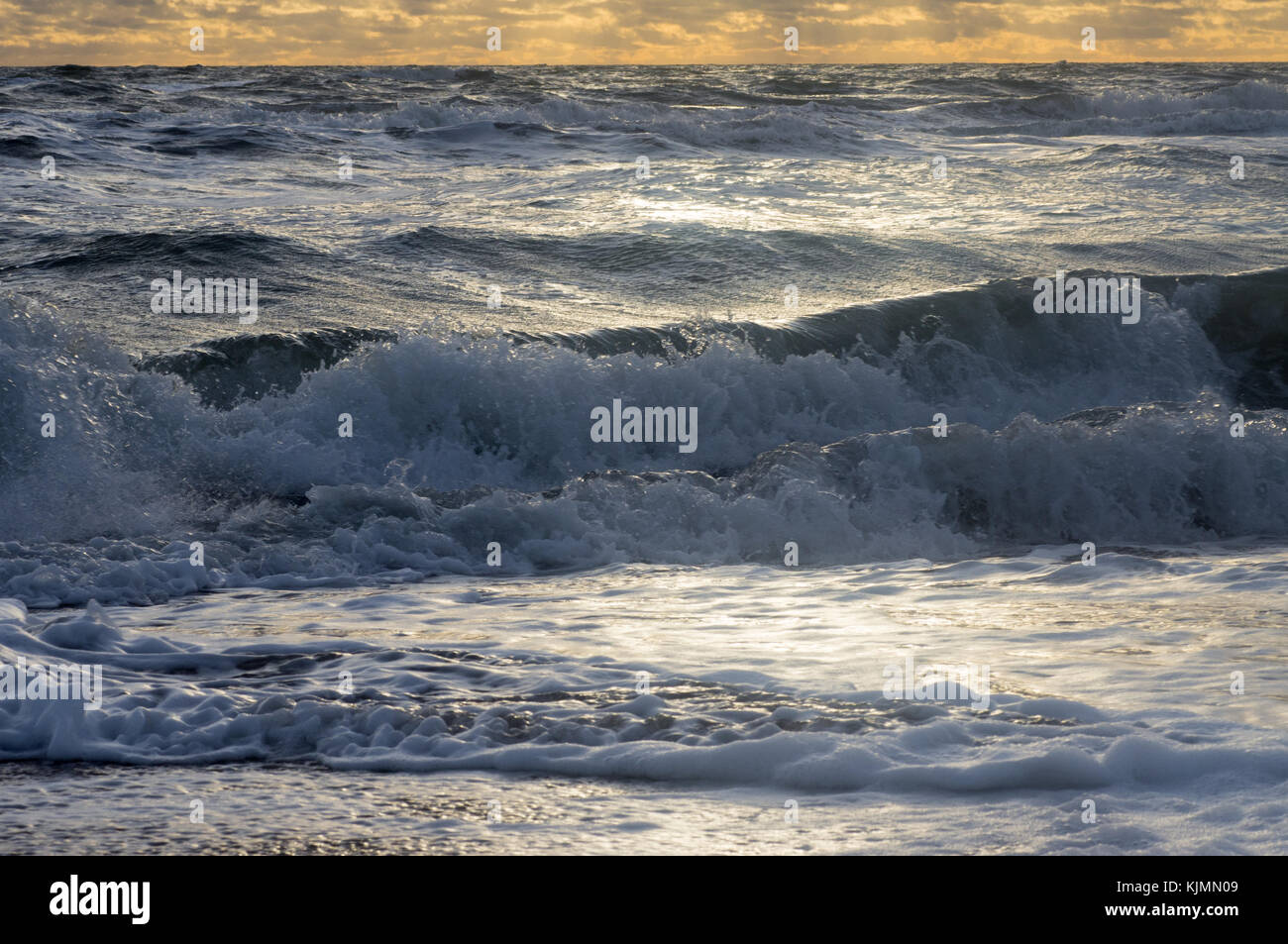Piccole onde in un mare in tempesta rotolare sulla riva con la schiuma e spray sotto il cielo nuvoloso e i raggi del sole Foto Stock