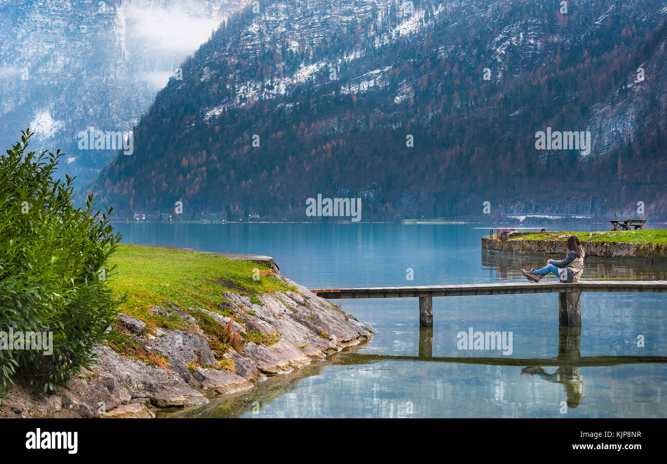 Giovane donna seduta su un ponte di legno, rilassante e ammirando le maestose montagne del Dachstein e il lago hallstatter, situato in hallstatt, Austria. Foto Stock