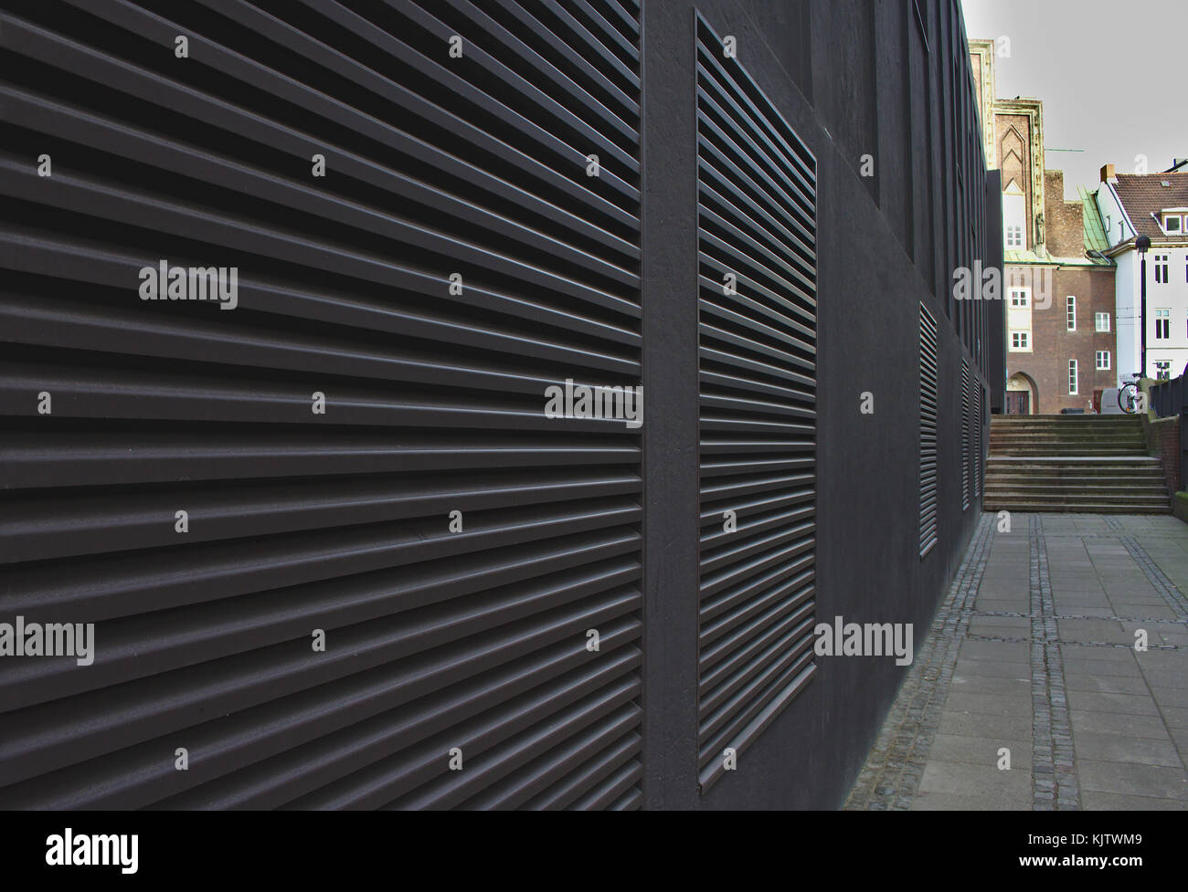 Marciapiede e parete nera con ampie bocchette di ventilazione che formano linee convergenti che conducono a passi distanti Foto Stock