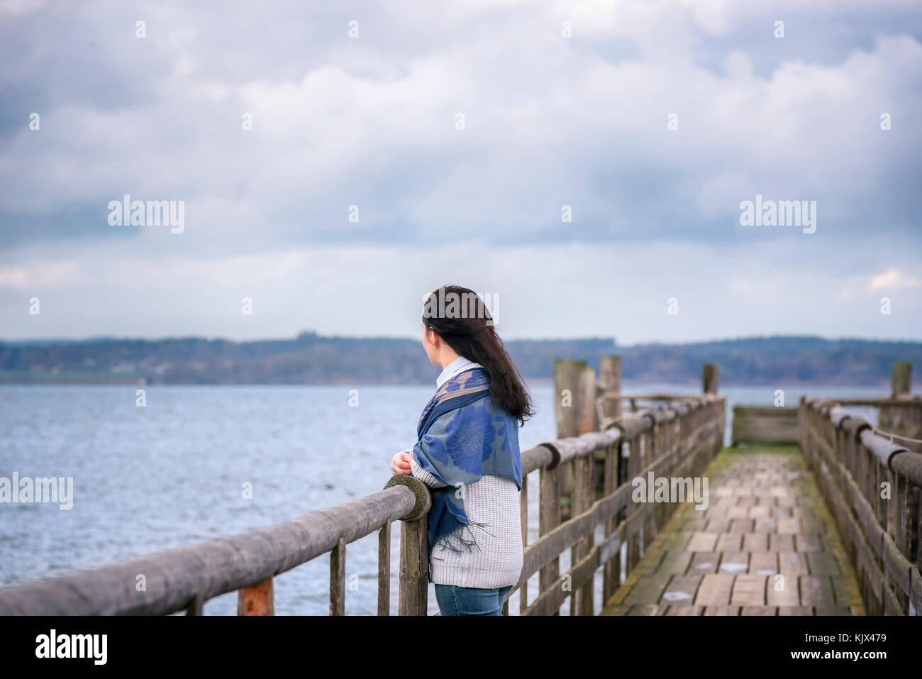Giovane donna in piedi da solo, su un vecchio ponte di legno, di pensare e di ammirare l'orizzonte nel pomeriggio, oltre il lago di Chiemsee, in Baviera, Germania. Foto Stock
